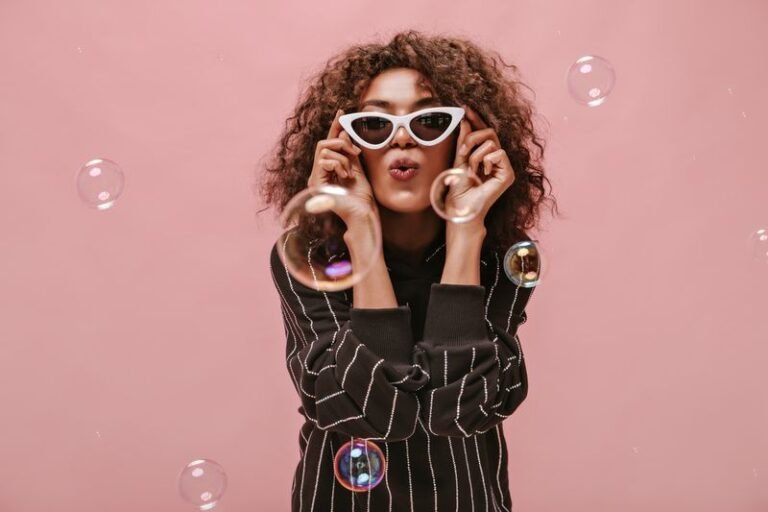 Stylish girl with short wavy hairstyle in striped black outfit blowing kiss, holding white sunglass